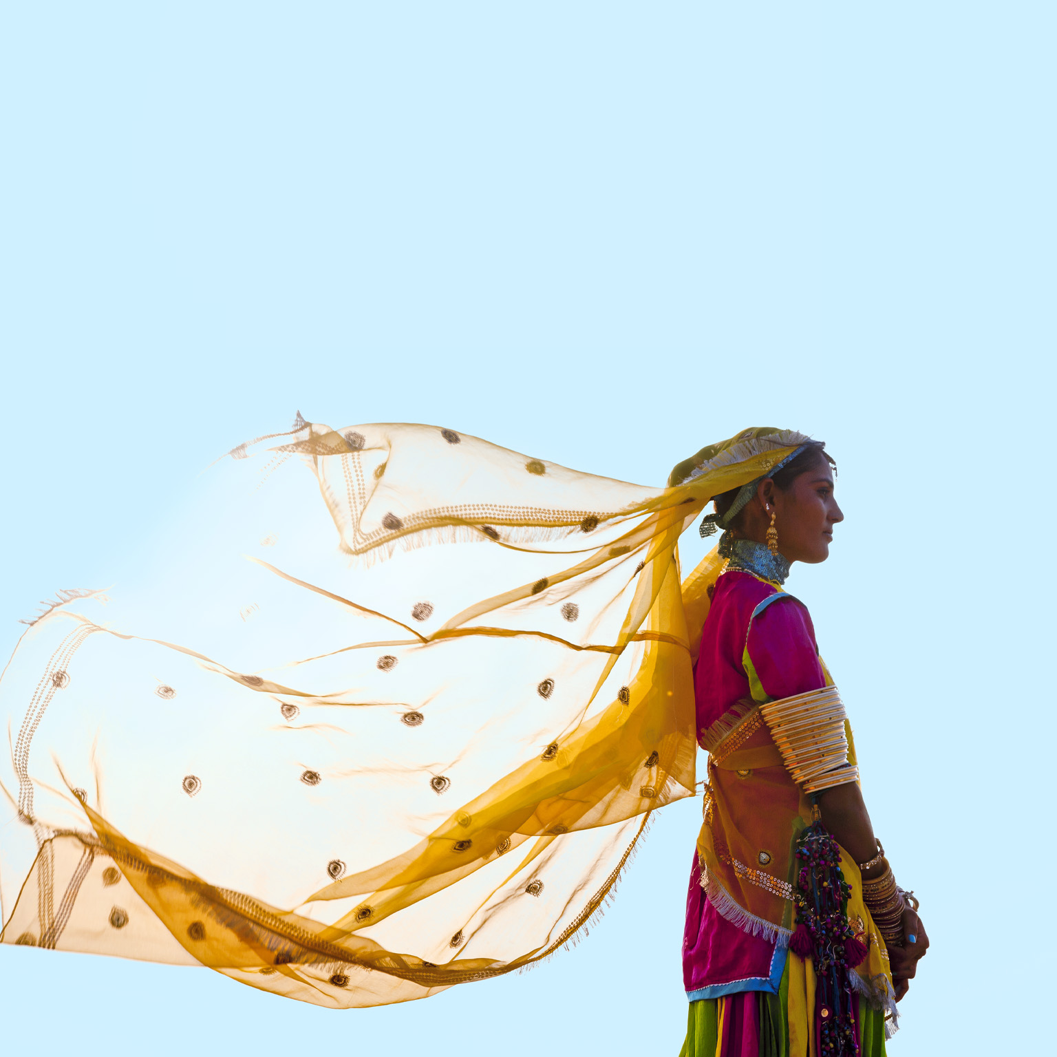A woman from India stands tall and proud in a vibrant saree, her yellow veil fluttering in the wind against a backdrop of a clear blue sky.