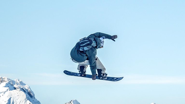 A snowboarder is captured midair performing a jump against a clear blue sky, with rugged, snow-covered mountains stretching across the background. The scene conveys motion and adventure, highlighting the contrast between the airborne rider and the vast alpine landscape below.