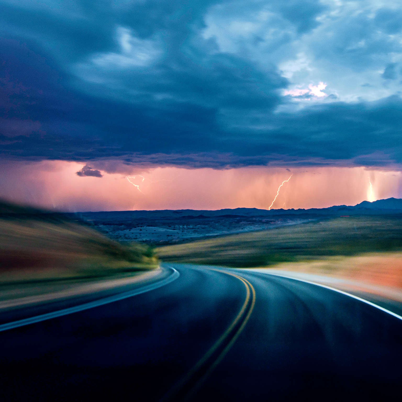 A dark asphalt road curves through a motion blurred landscape beneath a dramatic, stormy sky. In the distance, lightning flashes behind rain clouds, illuminating the horizon with an eerie light.