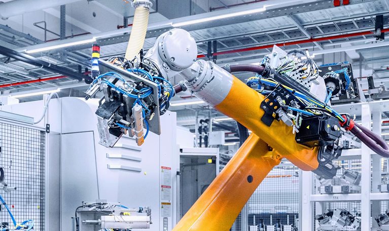 A large, bright industrial workspace with a prominent yellow robotic arm in the foreground. The robot appears to be working within an automated manufacturing system, surrounded by other machinery, parts, and storage racks.