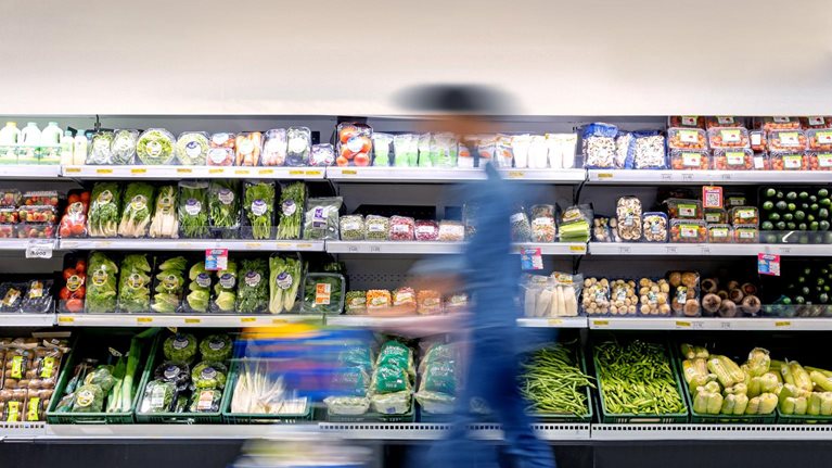 A man maneuvering a shopping cart through the grocery store, passing by the refrigerated produce section.