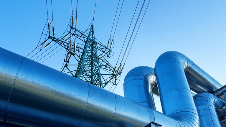 Large metal pipes tansporting oil curve and ascend against a clear blue sky with a tall electricity pylon visible in the background.