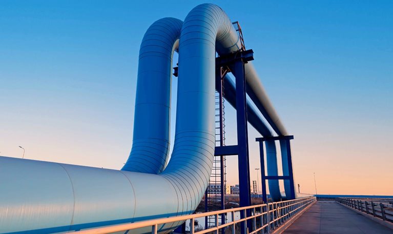 A large industrial pipeline rises and bends over a bridge-like structure, set against a clear blue sky at dusk. A paved walkway with railings runs alongside the pipes, leading the eye toward the horizon and giving the scene a clean, modern, and slightly futuristic feel.