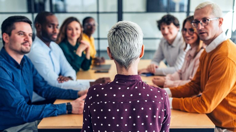 Image from behind of a woman with short gray hair, seated at a conference table, with a diverse group of professionals gathered around, looking at and intently listening to her.