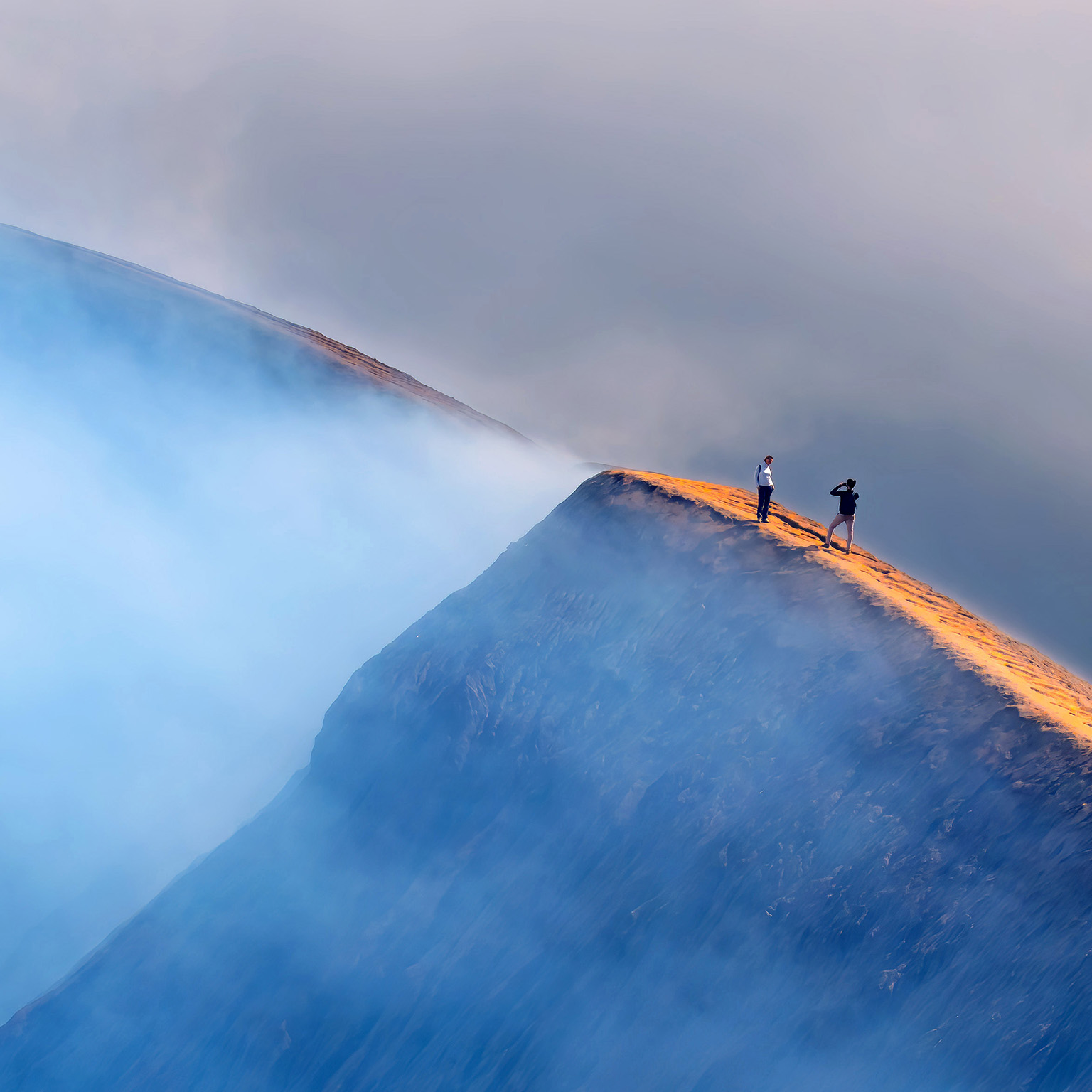 Aerial view of a mountain under clouds. Two walkers take photographs.