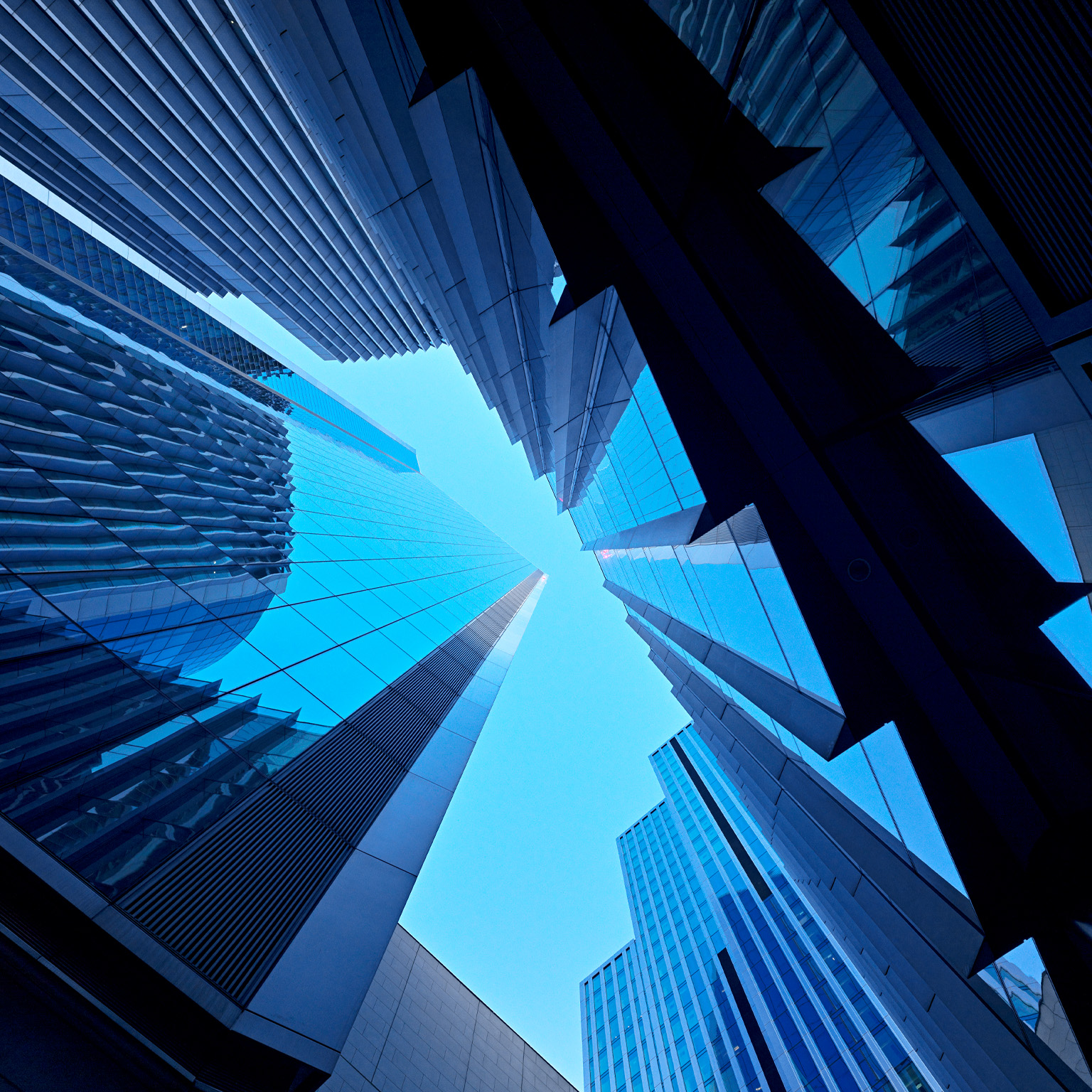 A dramatic upward view of modern glass skyscrapers converging toward the sky. The sharp lines and reflective surfaces create a sleek, futuristic feel.
