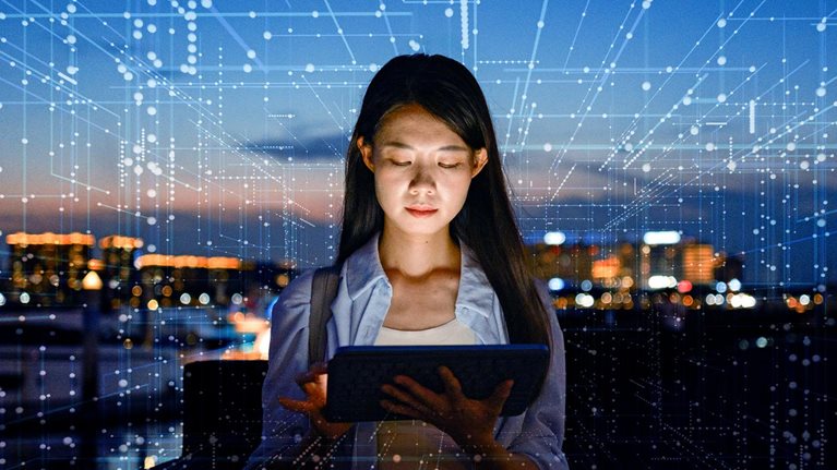 A woman stands outdoors at dusk, focused on a tablet in her hands with a city skyline glowing in the background. Digital lines and data-like patterns overlay the scene, suggesting connectivity, technology, and data flow.