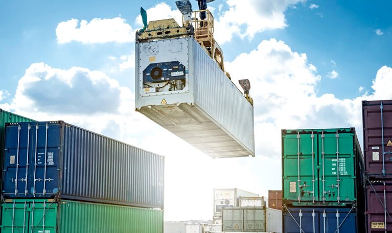 A large crane lifting a shipping container high above stacks of containers at a busy port. The bright sky and orderly rows of containers emphasize large-scale cargo handling and global trade operations.