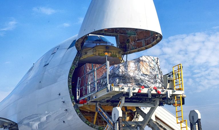 A cargo plane has its front end open as a sizable pallet is being loaded onto it.