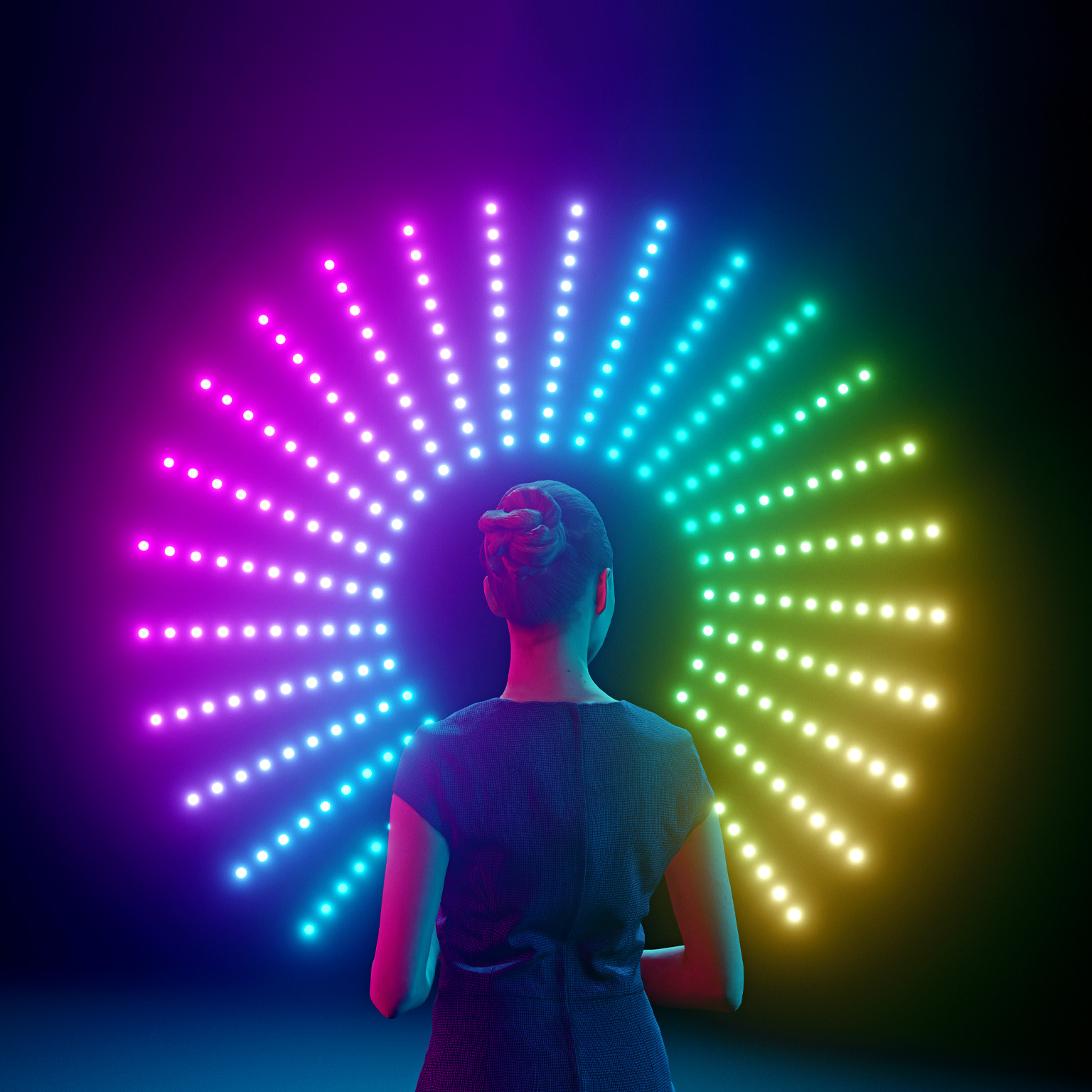 A woman with her back to the viewer stands in front of a circular arrangement of colorful lights. The lights are arranged in a rainbow pattern, transitioning from pink to blue to yellow.