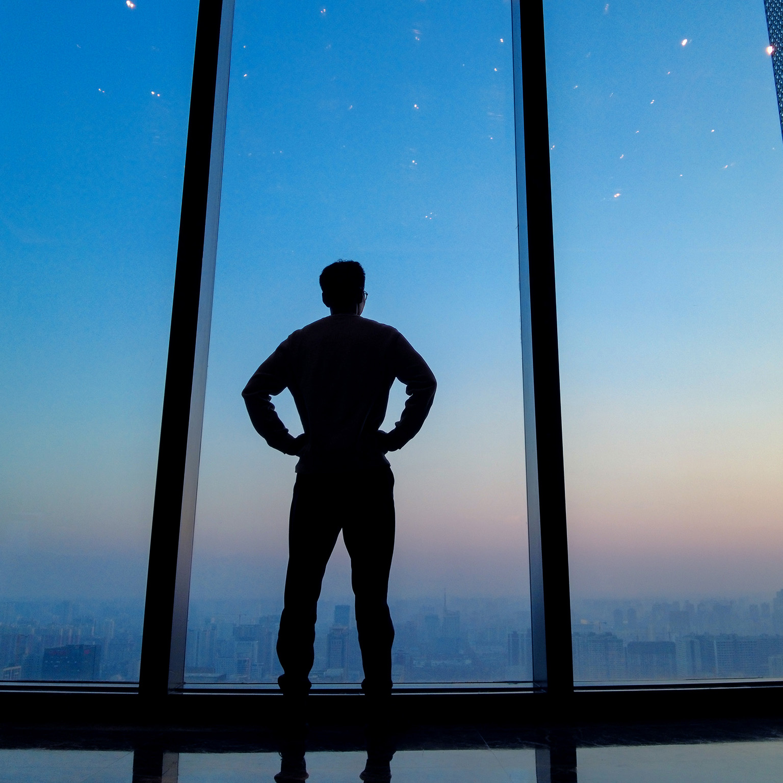 Young man staring out floor to ceiling office window looking over city skyline