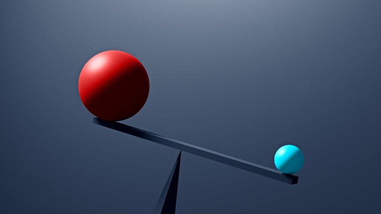 A large red sphere and a much smaller, light-blue sphere rest on opposite ends of a simple seesaw, which is unbalanced with the smaller blue sphere outweighing the red one. The scene is set against a dark, neutral background, emphasizing the contrast in size and weight between the two spheres.