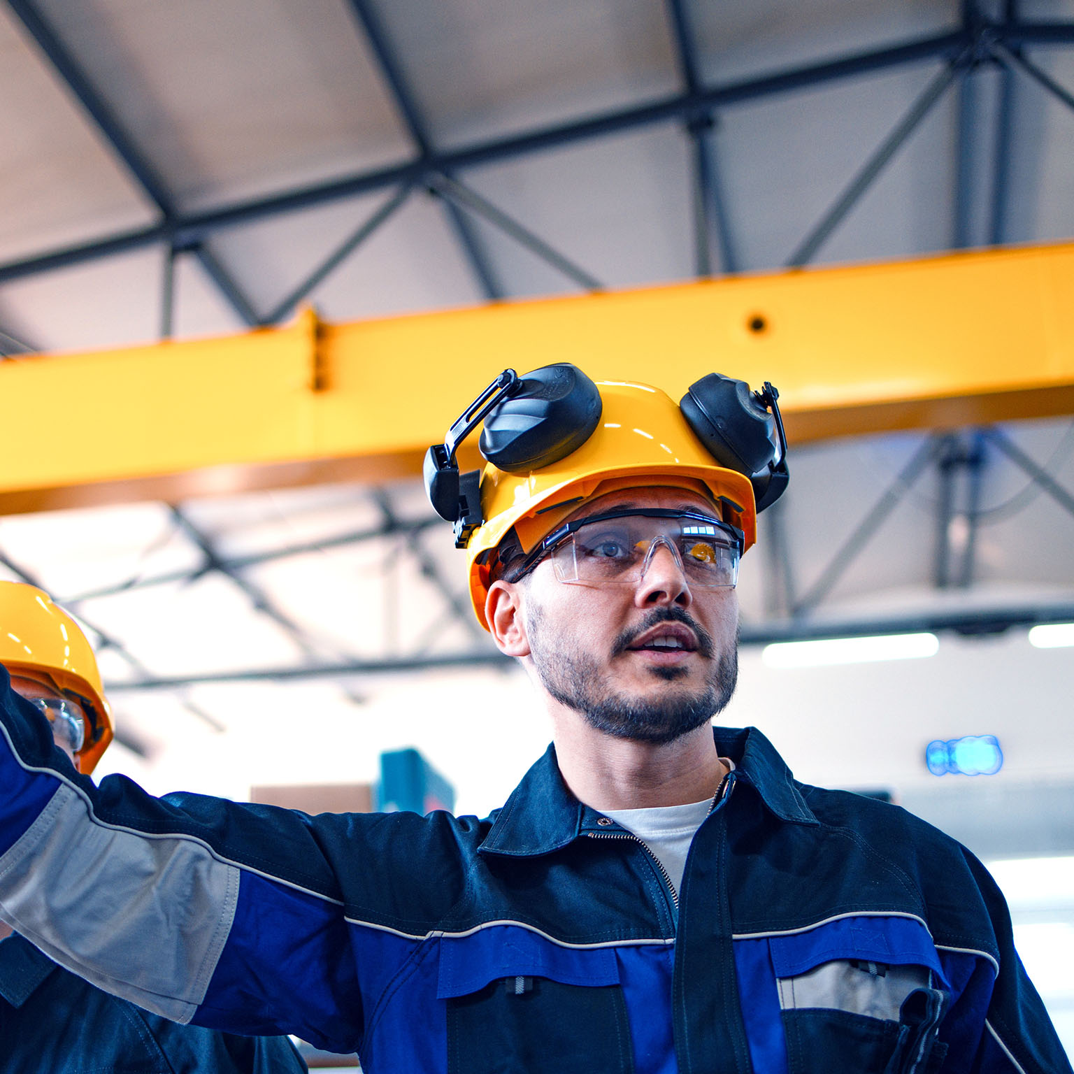 A focused male warehouse worker in safety gear inspects machinery with another worker, ensuring industry standards and safety protocols in a well-lit factory setting.