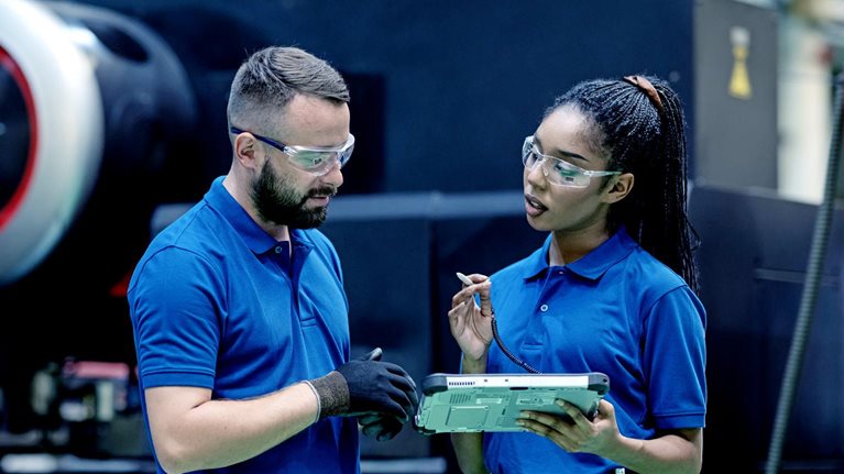 In an industrial manufacturing setting, a supervisor and process engineer, both dressed in blue company shirts and wearing protective glasses, engage in a conversation while examining a tablet near a sizable machine.