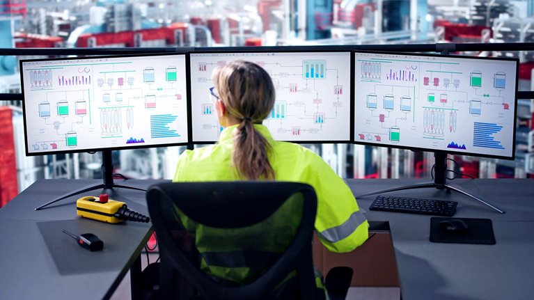 Image of an engineer operating an industrial data automation system using multiple computer screens with a factory in the background.