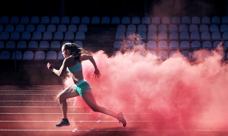 A determined female athlete races along a track, with a vibrant cloud of red smoke trailing behind her.