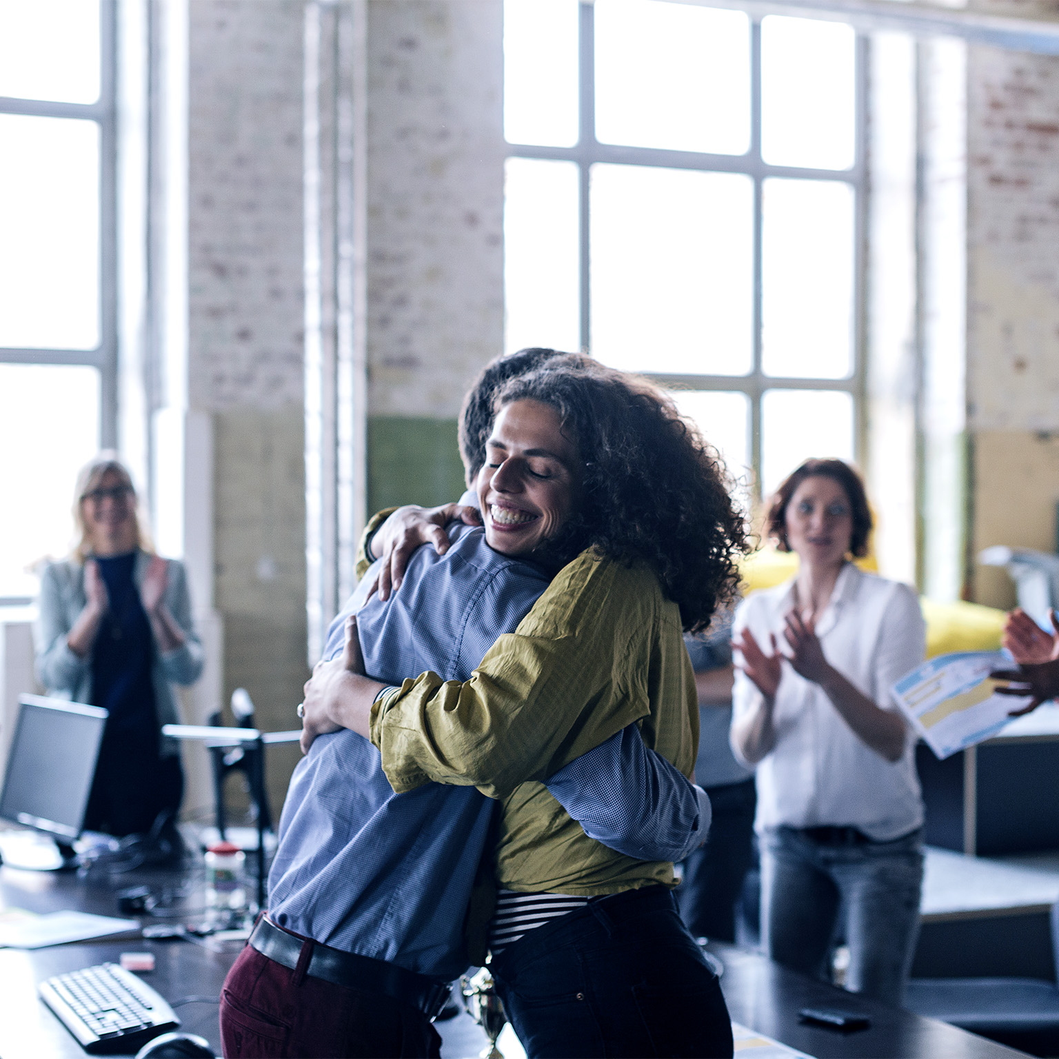 Image of people celebrating in an office with a woman smiling and being hugged by her coworker.