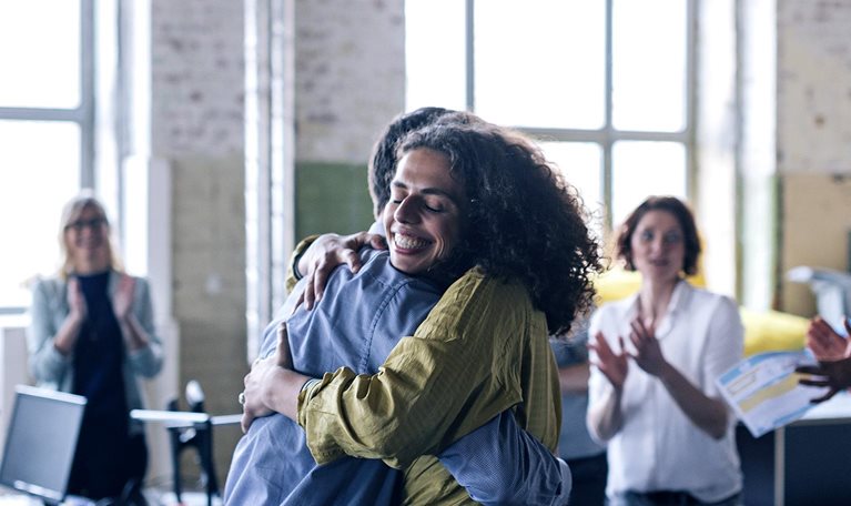 Image of people celebrating in an office with a woman smiling and being hugged by her coworker.
