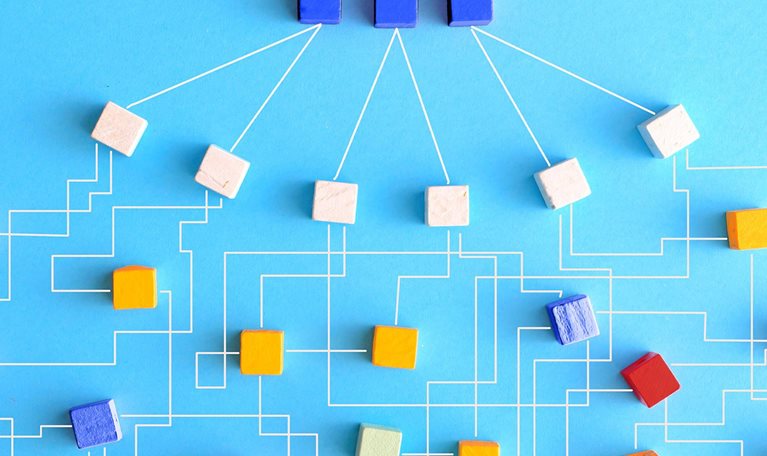 An organization chart constructed from wooden blocks that are linked together with delicate white lines. At the uppermost level of the chart, there are three blue blocks that connect to six white blocks. These white blocks then link outwards to form a larger network of blocks in varying shades of orange, red, and blue.
