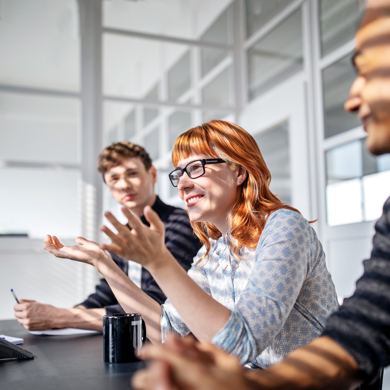 Three happy and engaged employees sitting at a board room table during a meeting. A woman sitting at the center of the group leans in to speak while gesturing expressively with her hands.  