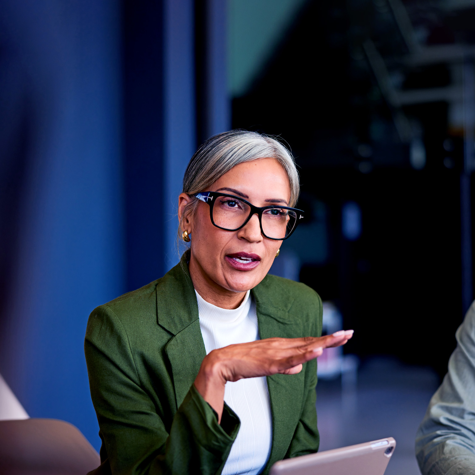 A mature businesswoman in a green blazer and glasses is speaking confidently while gesturing with her hands during a boardroom meeting.