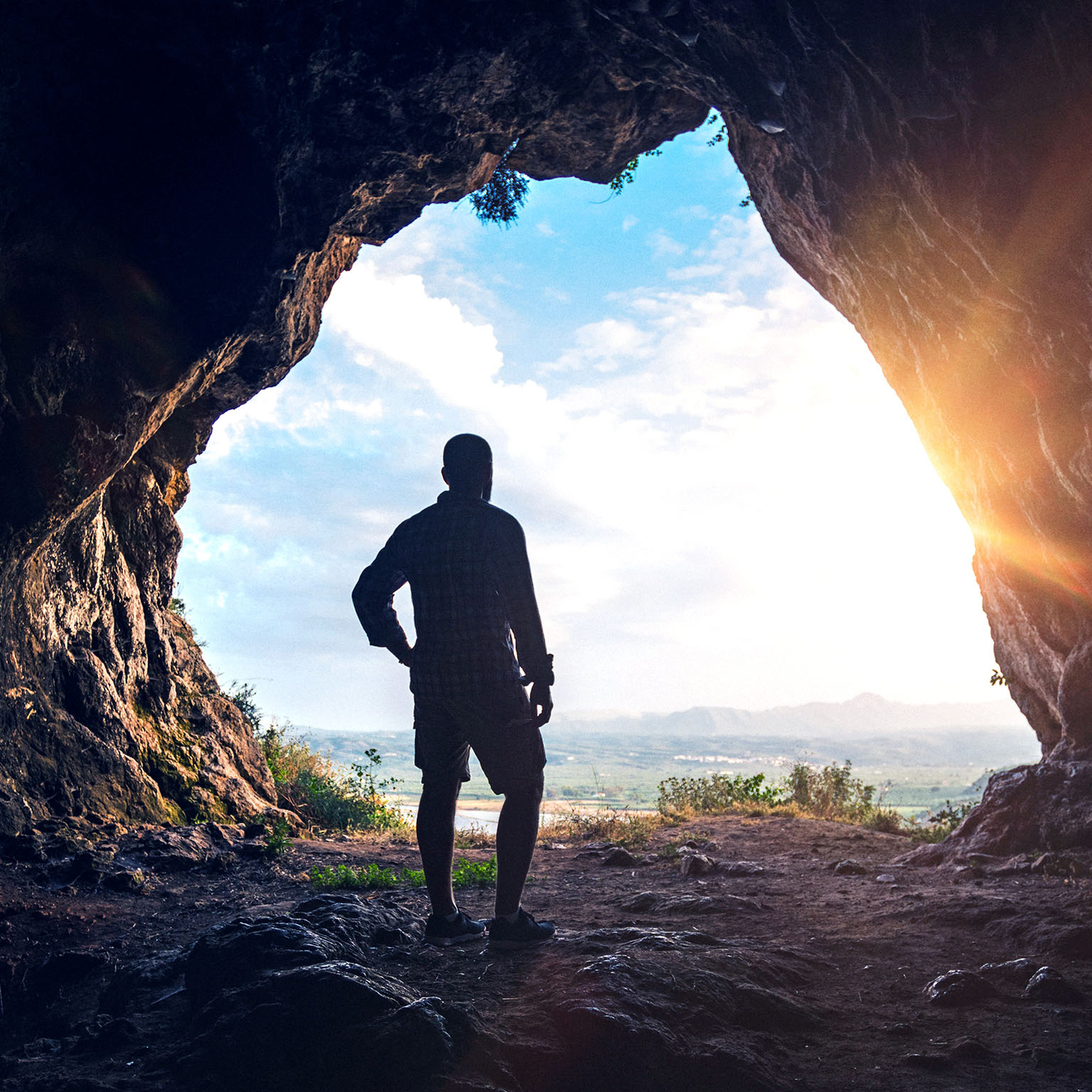 A hiker looks out over a sunset from inside the large opening of a cave