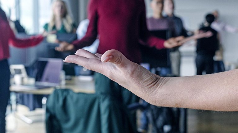 Close-up of a woman's upward facing hand in the foreground, with a blurred group of coworkers meditating in the background.