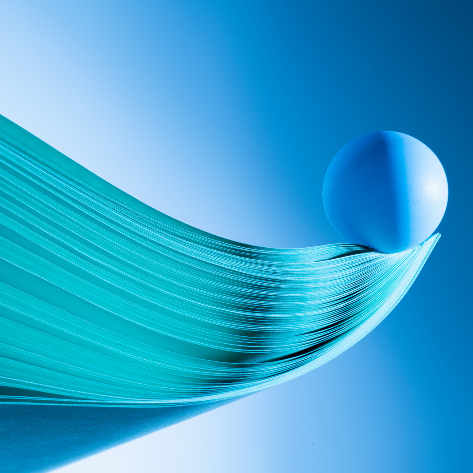  Close up of multiple sheets of green tinted paper in curve shape, blue ball balance at tip