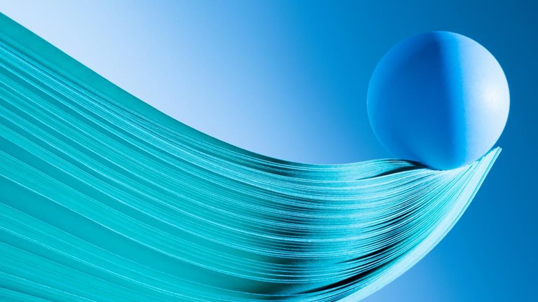 Close up of multiple sheets of green tinted paper in curve shape, blue ball balance at tip