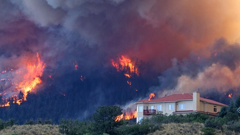 Image of a forest fire burning on a hillside, threatening houses below the blaze, with thick smoke and flames swirling and reaching for the sky.