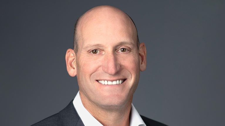 David Gitlin is standing against a neutral gray background, dressed in a dark suit jacket and white shirt, with a smile on his face.