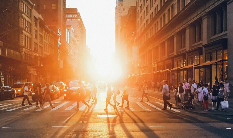 Sunlight shining on the diverse crowds of people walking across the busy intersection on 5th Avenue in New York City.