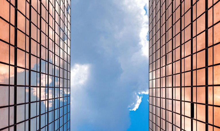 Looking up between two gleaming glass towers toward a vivid blue sky, the city reflects a bright future rising ahead.