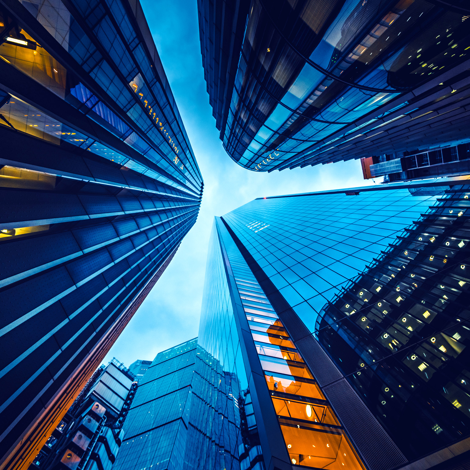 Looking up from street level at the skyscrapers in the heart of London's financial district. The buildings have glass facades and a moody blue cast from the setting sun. 