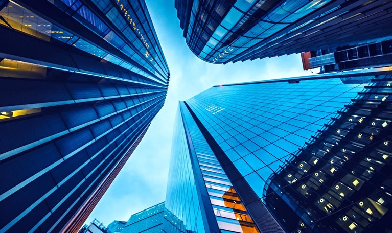 Looking up from street level at the skyscrapers in the heart of London's financial district. The buildings have glass facades and a moody blue cast from the setting sun.
