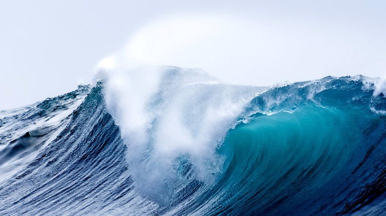 A large ocean wave is captured at its peak, with a deep blue color that transitions to vibrant turquoise as it curls over. The powerful wave is beginning to break, sending a spray of white foam high into the air against a pale sky.