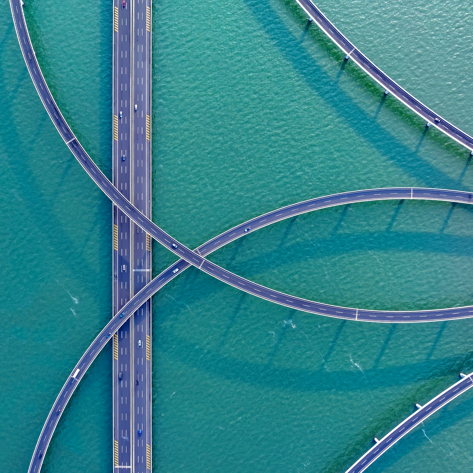 An aerial view of sweeping, elevated highways curving gracefully over turquoise water, creating an intricate, looping pattern. A few cars travel along the roads, adding scale and movement to the striking geometric scene.