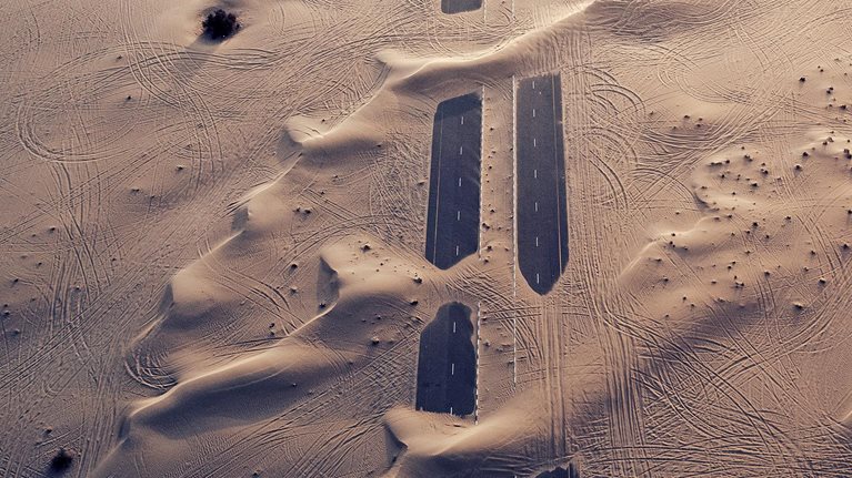 Sandy terrain with a road emerging in parts between stretches of sand.