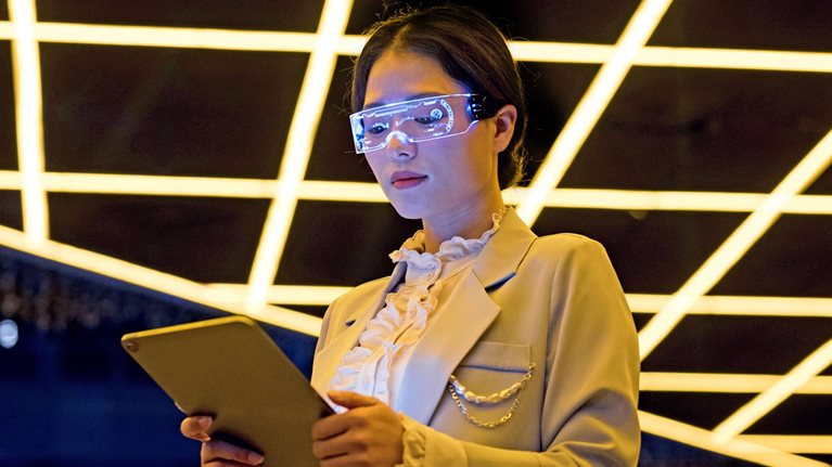 Image of a person wearing tech glasses and a lab coat looking at a tablet.