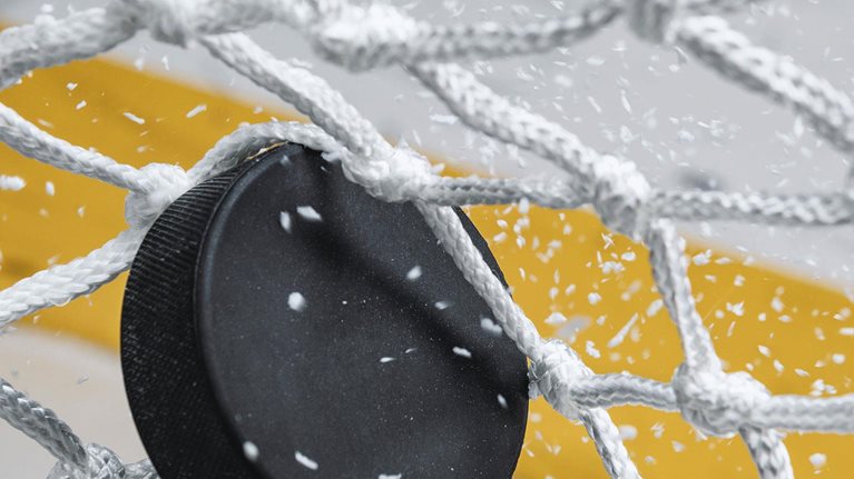 A close-up view of an Ice Hockey puck hitting the back of the goal net as shavings fly by, viewed from the front. Scoring a goal in ice hockey.