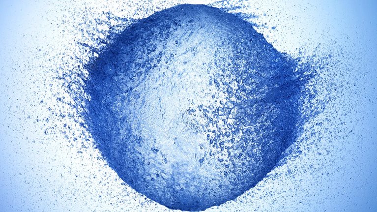 A high-speed photograph of a water balloon bursting. The water is flying outward in all directions, creating a roundish shape with a dynamic and explosive feel.