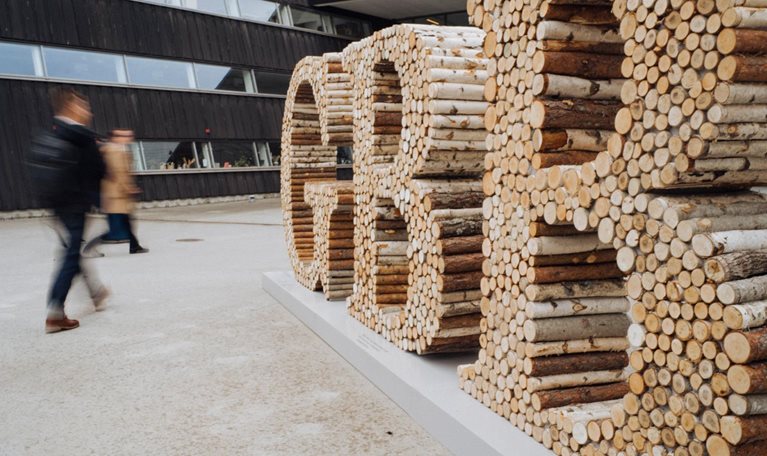 Blurred pedestrians walk past a large outdoor sculpture spelling “GBB,” constructed from stacked logs and branches, positioned in front of a modern building with dark wood cladding and long horizontal windows.
