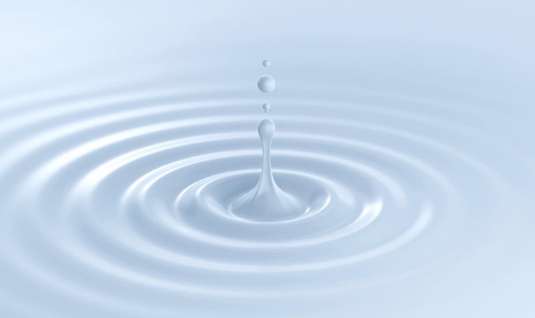 A single droplet of milky white liquid splashes up from a rippling surface. Gentle concentric ripples spread outward across the white background.