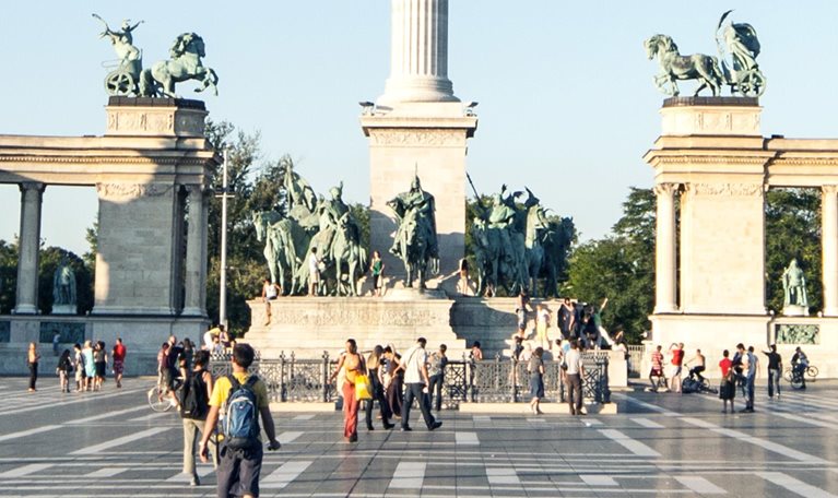 Visitors strolling and capturing images at Heroes Square in Budapest, with the Millennium column prominently displayed at the center of the view.