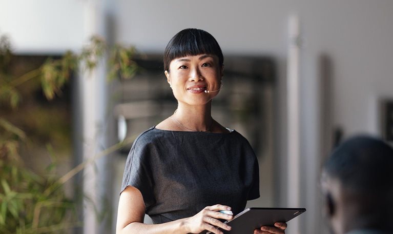 A confident presenter leads a business conference, engaging a diverse audience. The speaker is using a tablet and microphone, ensuring effective communication and interaction in a modern professional setting.