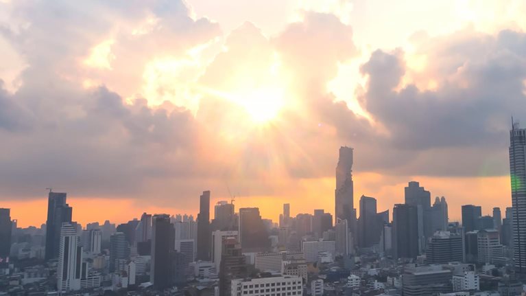 The image is an aerial view of the Bangkok skyline, likely near the Sathorn district, featuring several modern skyscrapers at sunset or sunrise. The sun is low on the horizon, casting a warm orange glow and dramatic light rays through the clouds.