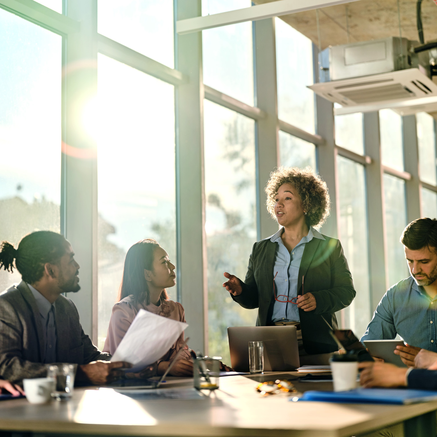  At the front of the conference room table, a woman is leading a meeting while a wall of large windows behind her allows light to stream in and cast long shadows over the table.