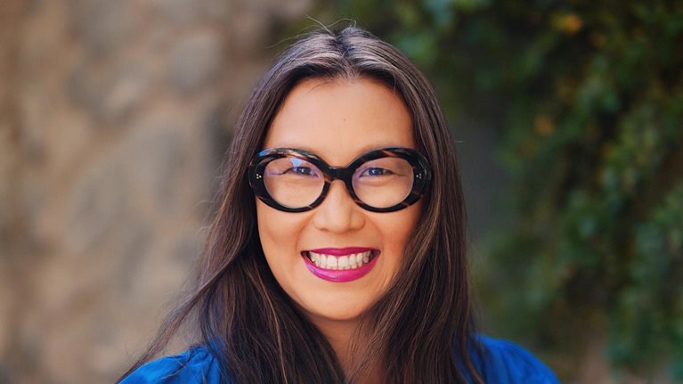 Portrait of a smiling Bonnie Wan with a stone wall and greenery blurred in the background.