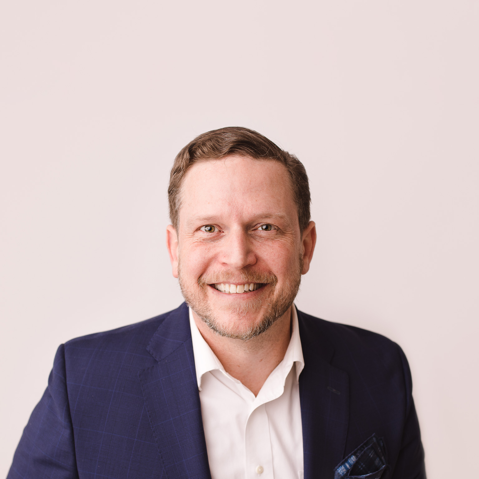 Nate Boaz, wearing a white button-down shirt and blue jacket, is captured with a smile on his face against a pale tan background