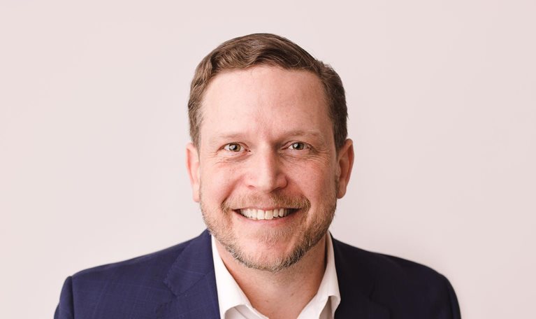 Nate Boaz, wearing a white button-down shirt and blue jacket, is captured with a smile on his face against a pale tan background